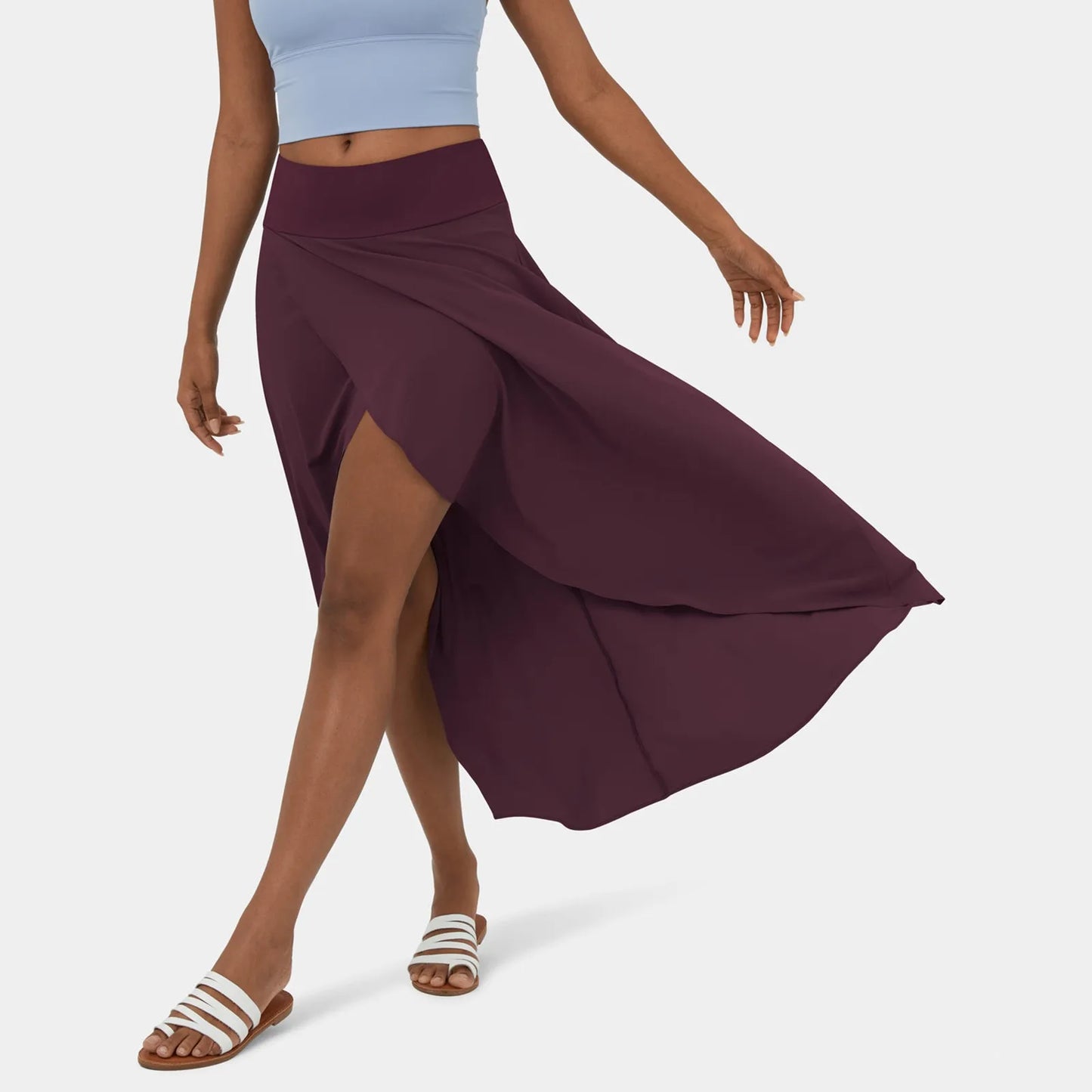 Een vrouw draagt een stijlvolle, donkerpaarse rok die elegant meebeweegt terwijl ze loopt. De rok heeft een hoge taille en een wijd uitlopende snit, die speels en luchtig beweegt. Ze draagt een lichtblauw sporttopje en witte sandalen, waardoor de outfit een frisse en casual uitstraling heeft.