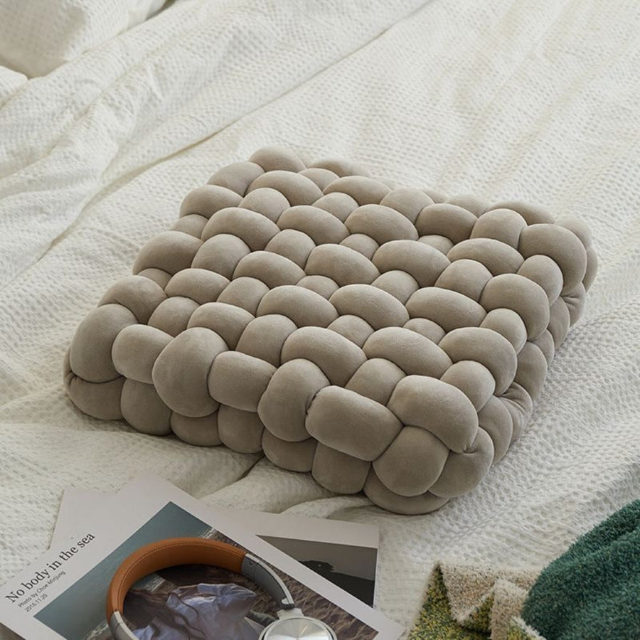 Elyisia comfort kussen in grijze tint, met een stijlvol gevlochten ontwerp, liggend op een bed. Het kussen biedt zachte ondersteuning en is ideaal als decoratief of zitkussen. Perfect om een verfrissende en moderne uitstraling aan je interieur toe te voegen.