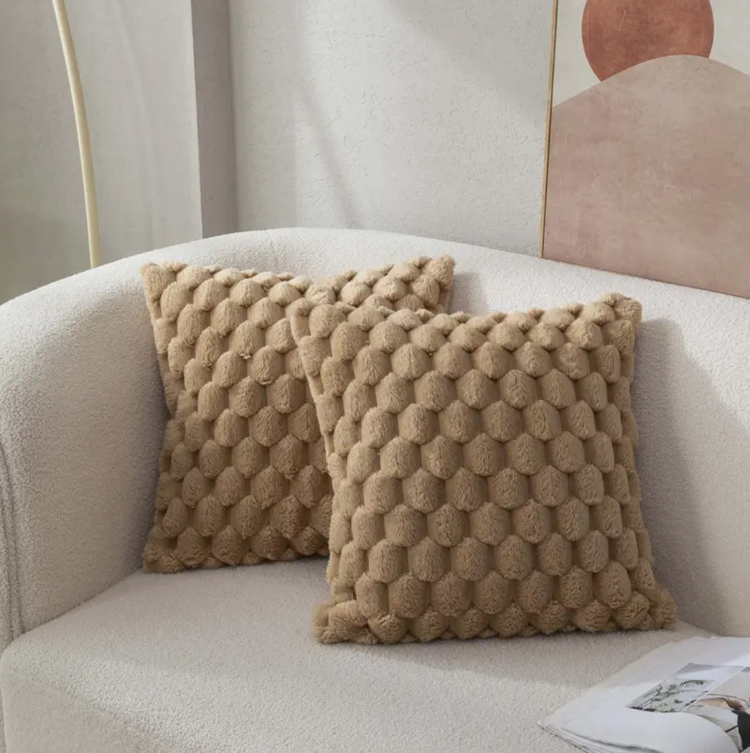 Twee beige, donsachtige kussens met een hexagonaal patroon liggen op een lichte, comfortabele bank, met een minimalistisch kunstwerk op de achtergrond.