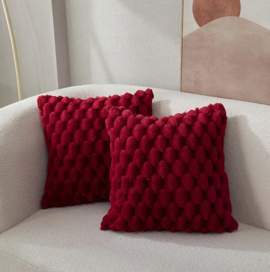 Twee diepe bordeauxrode, donsachtige kussens met een hexagonaal patroon liggen op een comfortabele, lichte bank, met een minimalistisch kunstwerk op de achtergrond.