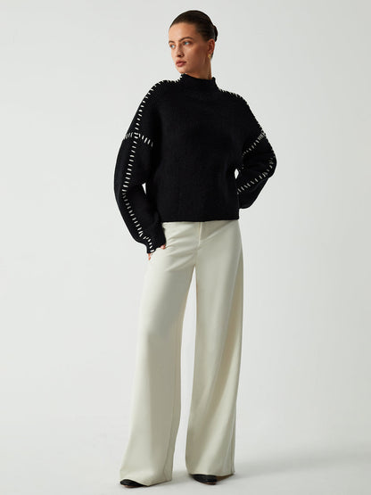 Model poseert in een Helvano oversized trui in zwart met witte stikseldetails langs de schouders en mouwen. De trui heeft een hoge kraag en een losse, comfortabele pasvorm. Gecombineerd met wijde, lichte broeken voor een moderne en elegante uitstraling. Perfect voor een casual maar stijlvolle look.