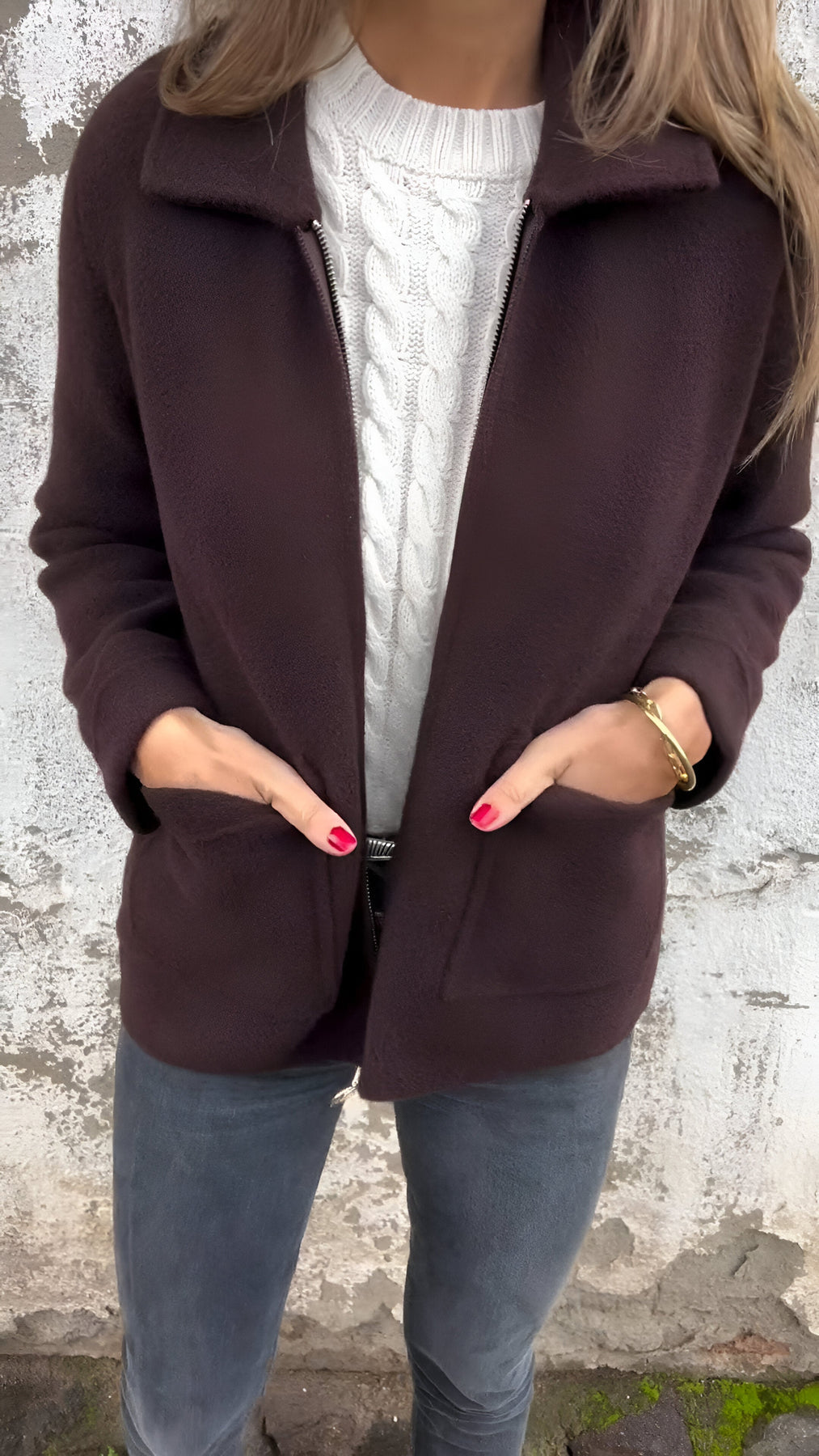 Kallia damesjas in donkerbruin, gedragen over een witte kabeltrui. De jas heeft een stijlvolle kraag, ruime zakken en een comfortabele pasvorm. Perfect voor een elegante en warme uitstraling in de winter.