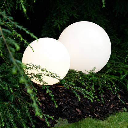 Twee witte sferische tuinlampen verlichten een groene tuin, gedeeltelijk bedekt door takken van bomen.