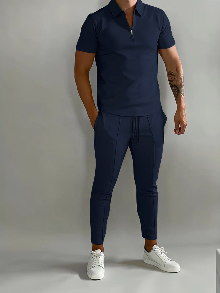 De afbeelding toont een man die een stijlvol, donkerblauw trainingspak draagt van het merk Opestico. Het trainingspak bestaat uit een korte rits polo en een bijpassende broek met trekkoord voor een comfortabele pasvorm. De man combineert het met witte sneakers, wat zorgt voor een sportieve, maar toch elegante uitstraling. Dit trainingspak is zowel comfortabel als modieus, ideaal voor dagelijks gebruik of een actieve levensstijl.