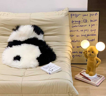 Een zwart-wit panda-vormig vloerkleed ligt op een beige bank, naast een stapel boeken en een schattige lamp in de vorm van een geel teddybeer. Het zachte, harige kleed biedt bescherming aan de bank en voegt een speelse, gezellige sfeer toe aan het interieur. Het unieke ontwerp van het vloerkleed maakt het een charmant accessoire in de woonkamer.
